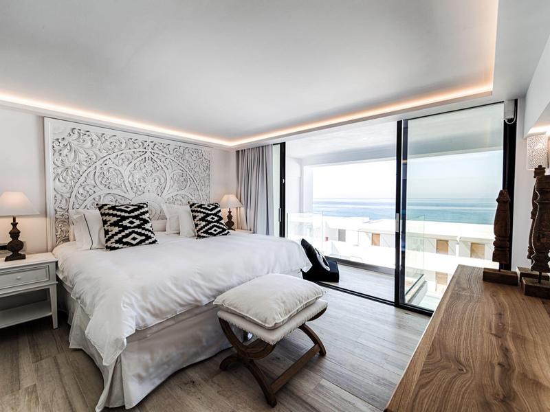 Moderne slaapkamer met groot bed, decoratieve kussens, houten meubels en balkon met oceaanzicht.