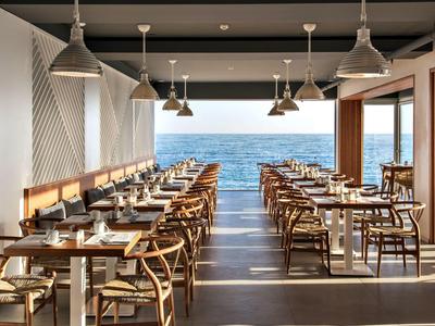 Modern restaurant met zeezicht en houten stoelen, daglicht stroomt binnen.