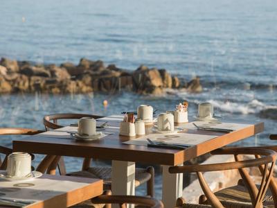 Tavoli apparecchiati con tazze e caffettiere su una terrazza sul mare con rocce sullo sfondo.