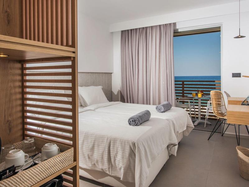 Licht hotelkamer met twee eenpersoonsbedden, bureau en balkon met uitzicht op zee.