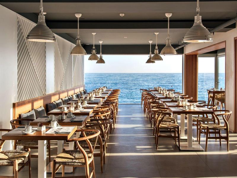 Modern restaurant met zeezicht en houten stoelen, daglicht stroomt binnen.