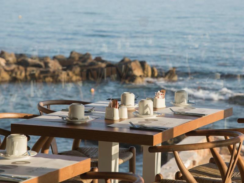 Tafels met kopjes en koffiepotten op een terras aan zee met rotsen op de achtergrond.