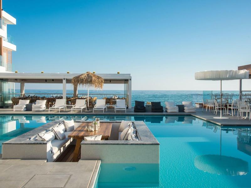 Modern hotelzwembad met loungegedeeltes, ligbedden en zeezicht bij heldere lucht.