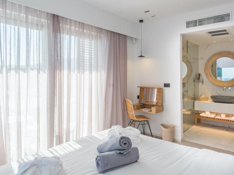 Lichte hotelkamer met groot raam, bureau en moderne badkamer met spiegel.