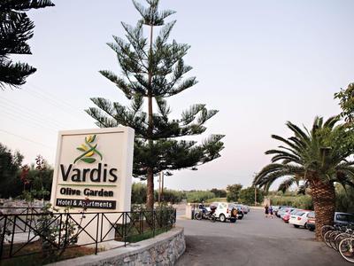 Vstup do Vardis Olive Garden s stromy a zaparkovanými auty podél silnice.