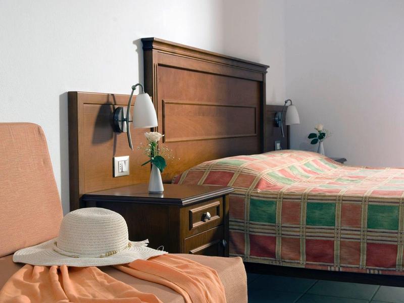 Ein schlichtes Hotelzimmer mit einem Bett in buntem Karo-Design und einem Stuhl mit Hut.