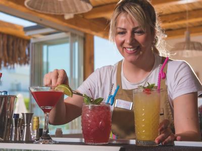 Frau bereitet bunte Cocktails an einer Bar mit Holzelementen im Hintergrund zu