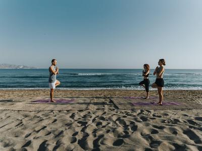 Drei Personen machen Yoga am Strand mit Blick auf das Meer unter klarem Himmel.
