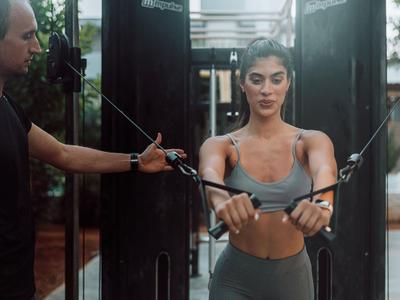 Frau trainiert an einem Kabelzug im Fitnessstudio, geführt von einem Trainer.