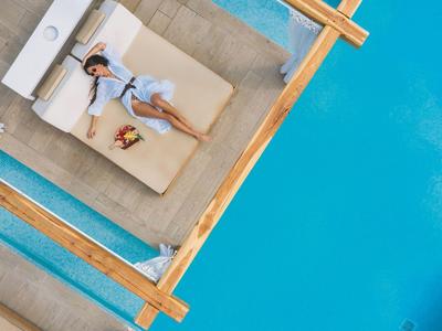 Frau mit Hut und Sommerkleid entspannt sich auf einer Sonnenliege neben einem blauen Pool.
