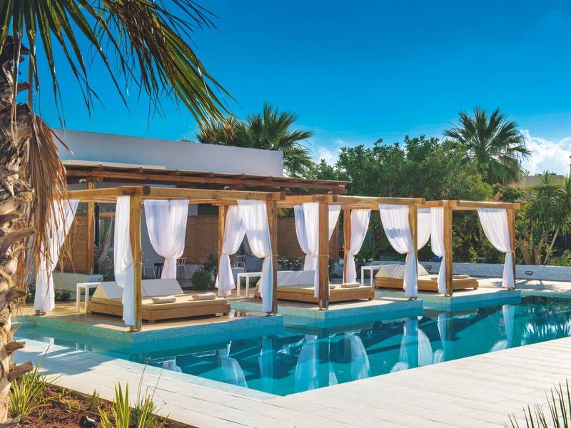 Piscina all'aperto con lettini sotto pergolati e palme vicino a un hotel.