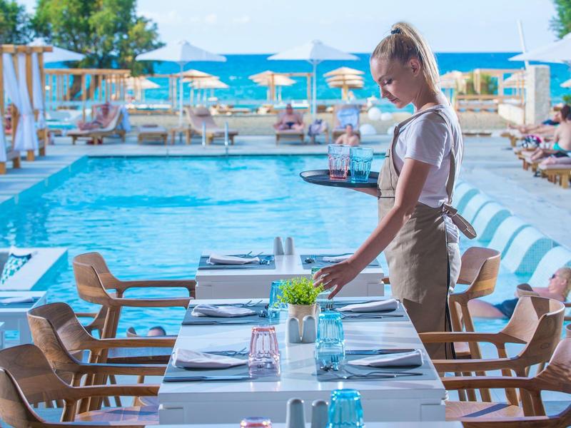 Donna che sistema un tavolo apparecchiato vicino a una piscina in una struttura balneare soleggiata.