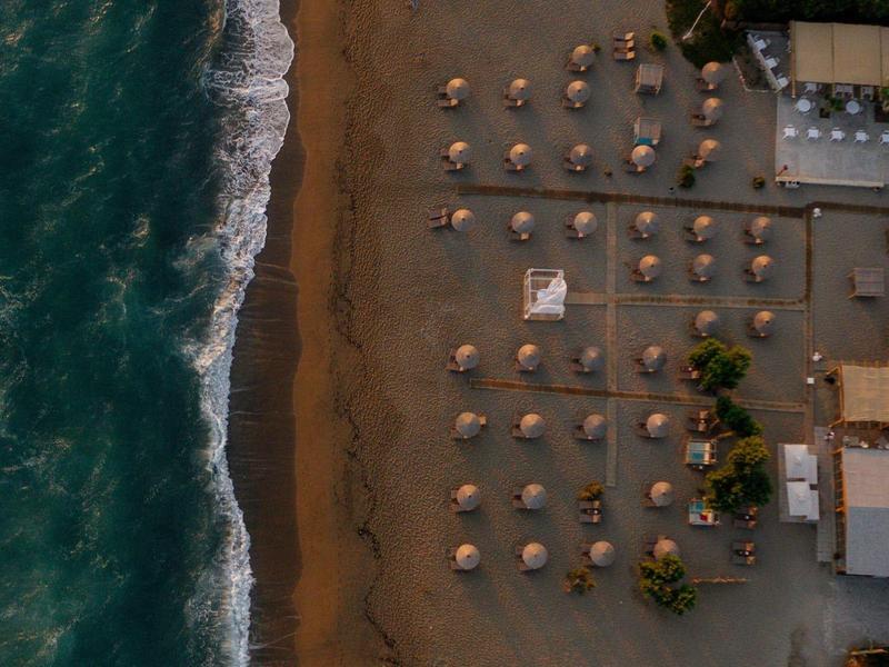 Veduta aerea di una spiaggia con ombrelloni di paglia e onde che si infrangono.