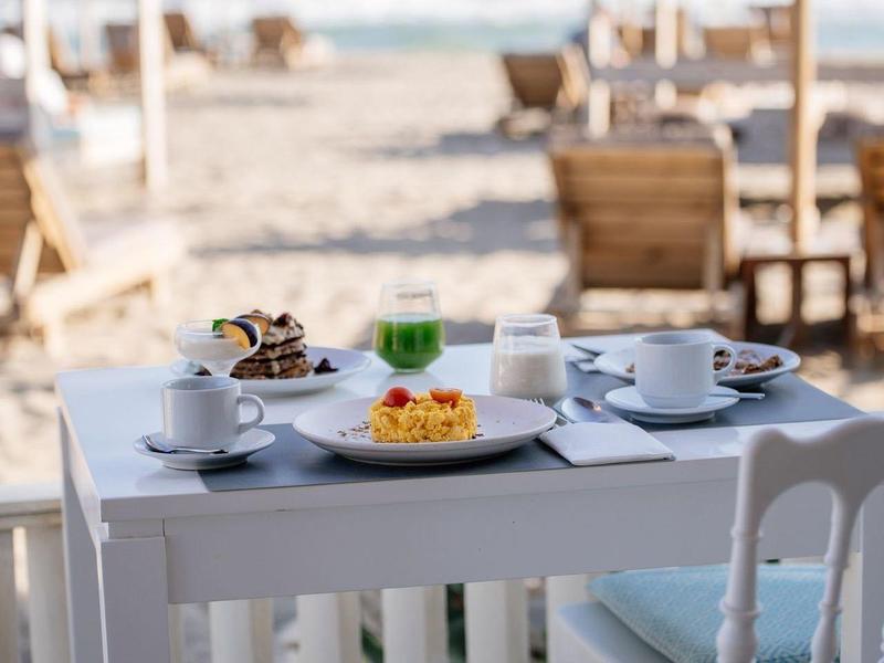 Tavolo bianco con colazione davanti al mare su una spiaggia con ombrelloni.