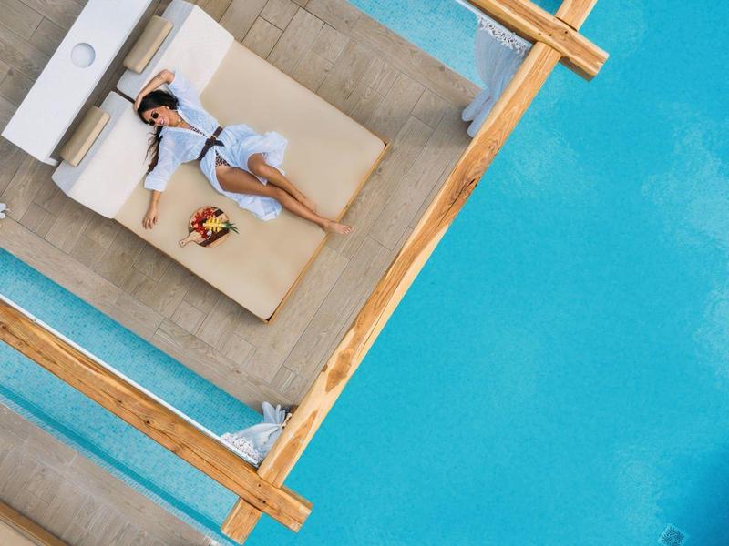 Una donna in abito bianco rilassata su un divano beige accanto a una piscina azzurra.