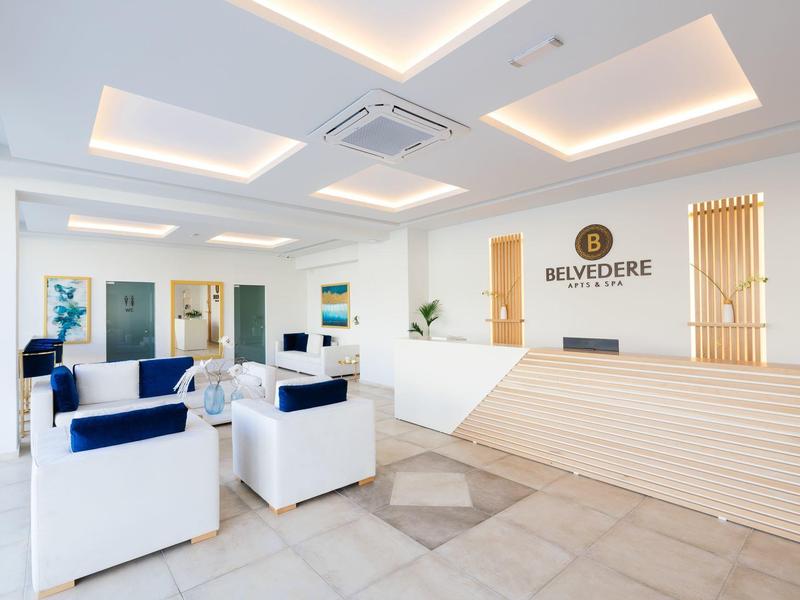 Světlá hotelová recepce s moderní dřevěnou recepční přepážkou a bílými křesly s modrými polštáři