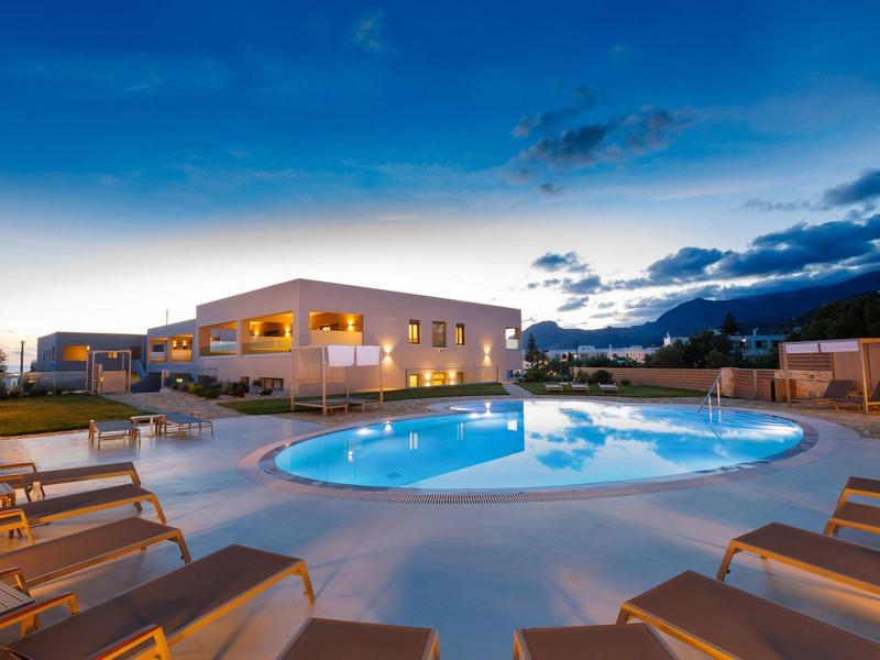 Večerní snímek osvětleného bazénu s lehátky před moderní hotelovou budovou.