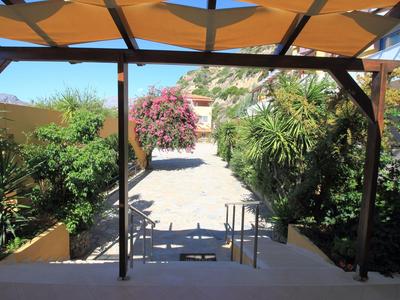 Vue depuis une terrasse couverte sur un chemin bordé de plantes méditerranéennes et une maison sur une colline.
