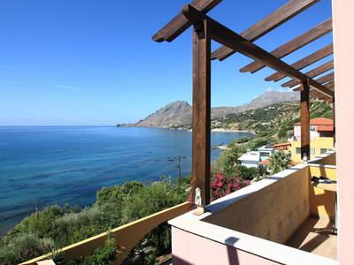 Balkon mit Holzdach und Blick auf das blaue Meer und Hügel bei klarem Himmel.