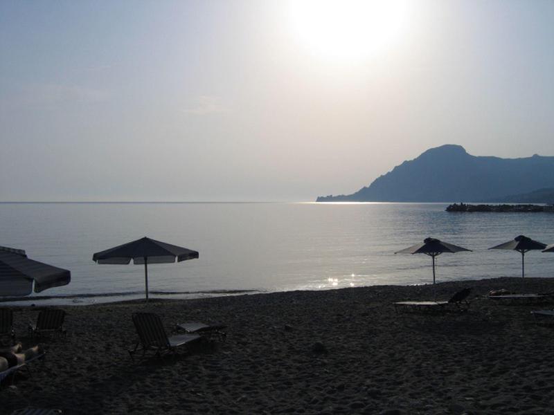 Strand met ligstoelen en parasols bij kalme zee en bergzicht in zonlicht.