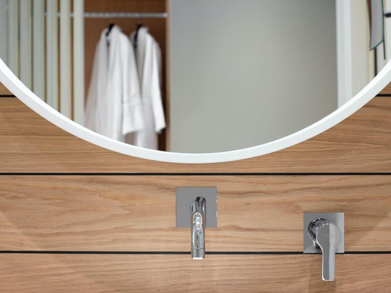 Nowoczesne baterie łazienkowe na drewnianej obudowie z lustrzanym odbiciem i białymi szlafrokami w tle.