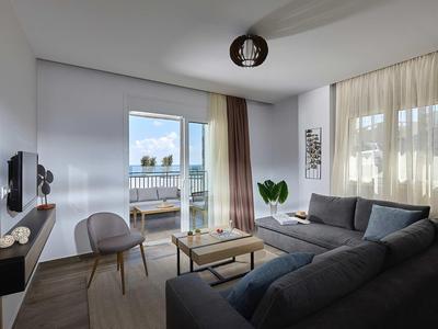 Jasny salon z dużymi oknami, szarą sofą i widokiem na wodę i miasto.