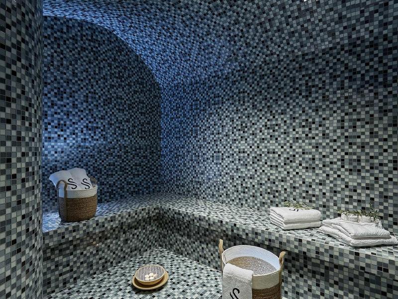 Nowoczesna łaźnia parowa z mozaikowymi płytkami w chłodnych odcieniach niebieskiego i białymi ręcznikami.