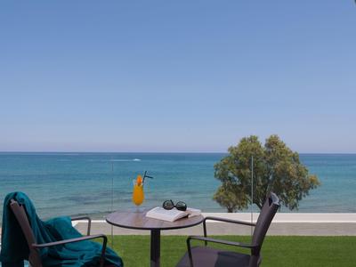 Blick auf einen Tisch mit Getränk, Stühlen und Baum vor blauem Meer und klarem Himmel.