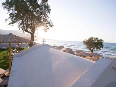 Strand mit weißen Gebäuden, Bäumen und Sonnenschirmen am Meer bei Sonnenuntergang.