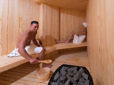 Zwei Männer entspannen in einer hellen, holzverkleideten Sauna mit Saunaofen und Holzeimer.