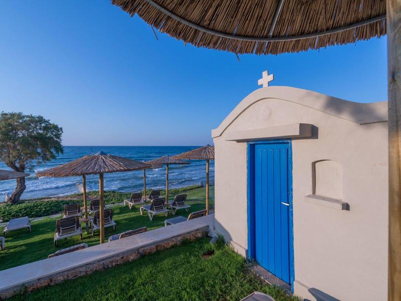 Klein wit gebouw met blauwe deur dicht bij strand met parasols en een boom onder een heldere blauwe lucht