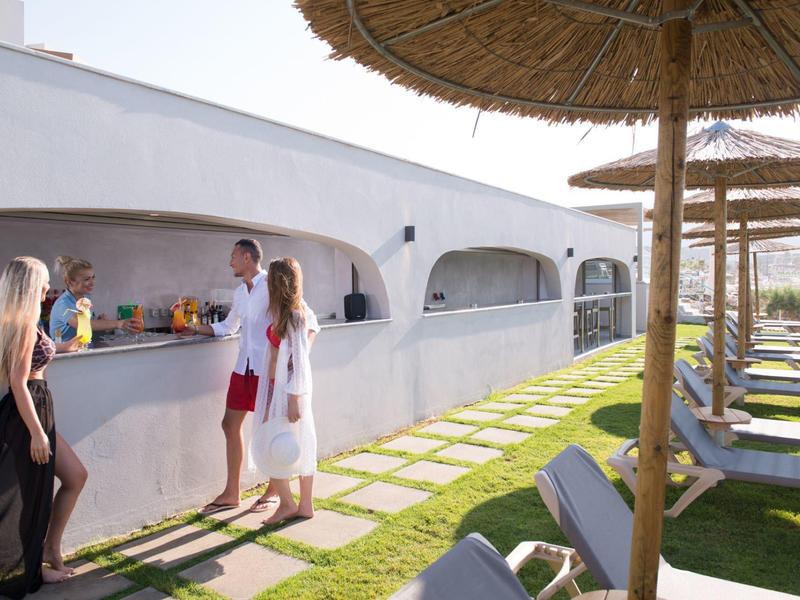 Mensen kletsen bij een witte gebogen hotelbar met zwembadligstoelen en parasols in de buurt.