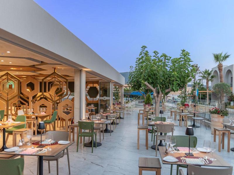 Moderne buitenruimte van een restaurant met tafels, stoelen en bomen onder een blauwe lucht.