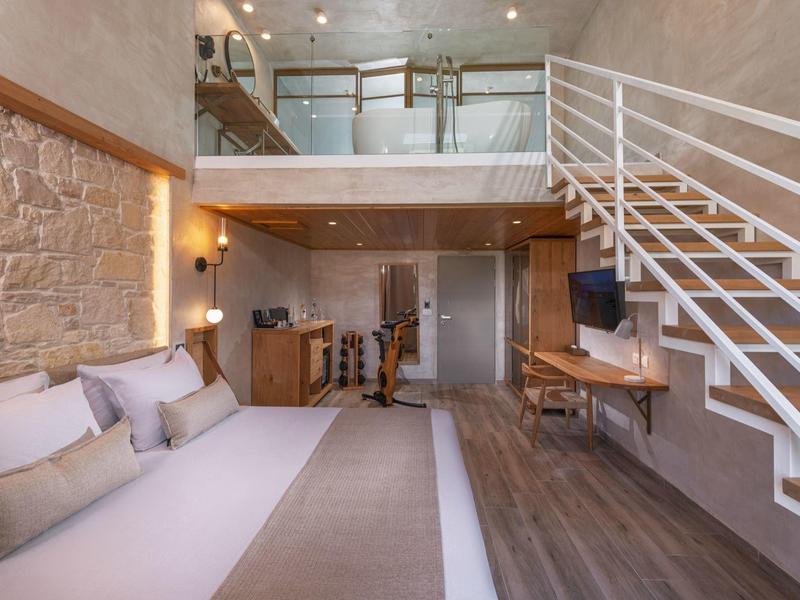 Moderne hotelkamer met groot bed, houten vloer, stenen muur, trap naar vide en TV.