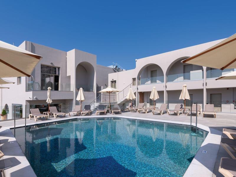 Rond zwembad omgeven door witte gebouwen en parasols onder een heldere lucht.