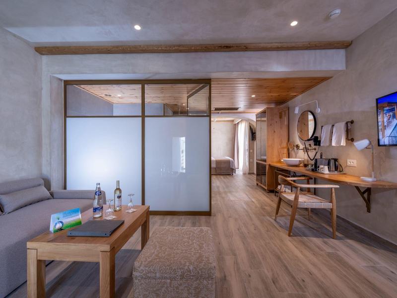 Moderne hotelsuite met houten vloer, grijze bank, glazen afscheiding en bureau met stoel en tv.