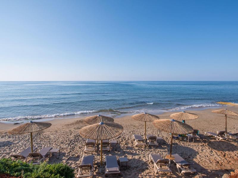 Zonnig strand met strooien parasols en ligbedden aan rustige zee onder heldere blauwe lucht.