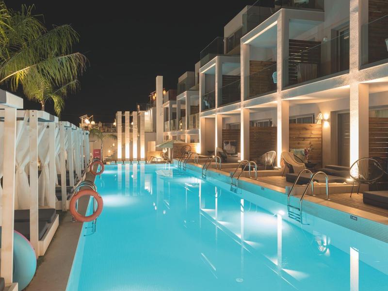 Oświetlony basen na zewnątrz z leżakami i nowoczesnymi pokojami hotelowymi nocą.