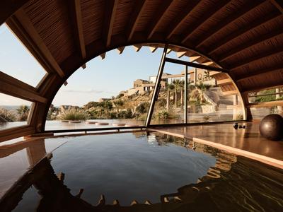 Vista desde una piscina cubierta hacia un paisaje costero mediterráneo soleado con casas.