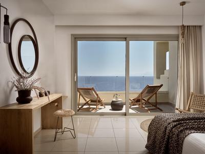 Camera d'hotel luminosa con vista mare, due sedie a sdraio sul balcone, scrivania in legno e specchio rotondo.