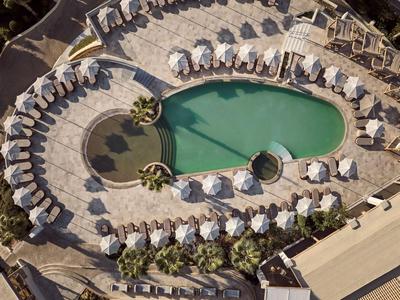 Veduta aerea di una piscina curva circondata da ombrelloni e lettini in un hotel.