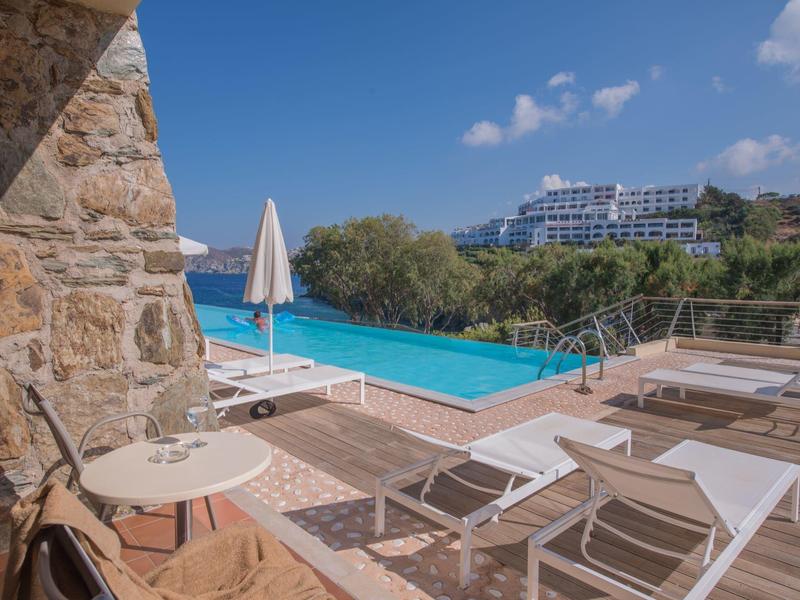 Piscina a sfioro con lettini e hotel su una collina sotto un cielo azzurro.