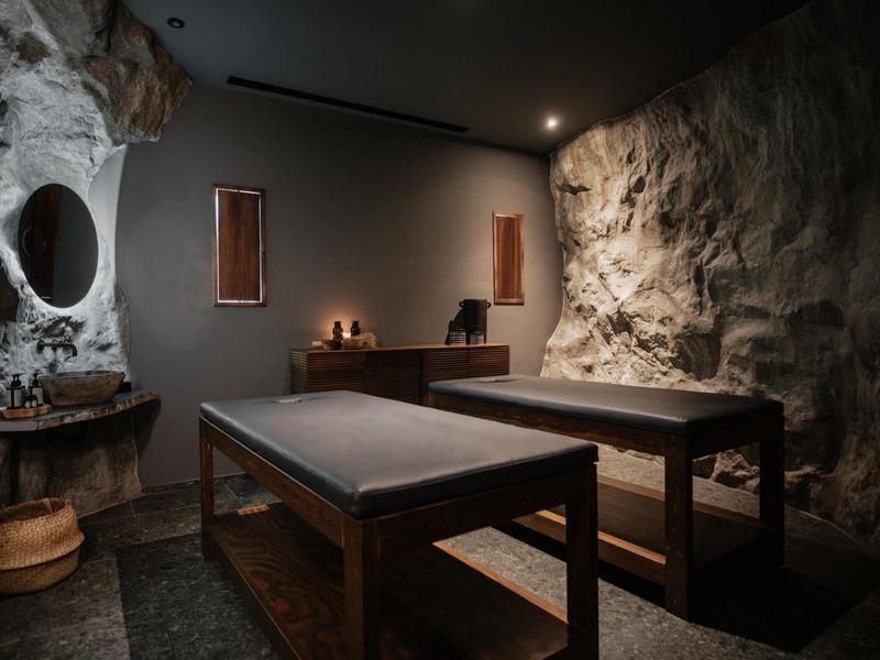 Accogliente stanza spa con tavoli da massaggio in legno e illuminazione soffusa in un ambiente con parete in pietra rustica.