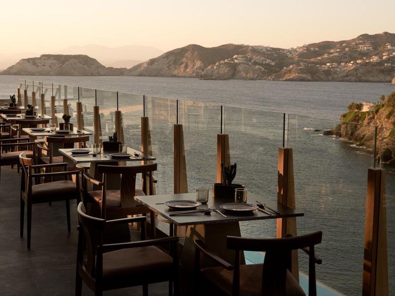 Ristorante all'aperto con vista sul mare e costa montuosa al tramonto.