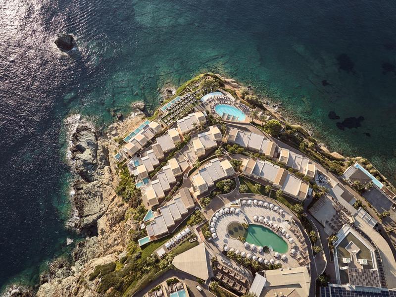 Vista aerea di un resort costiero con piscine, circondato da mare turchese limpido e costa rocciosa.