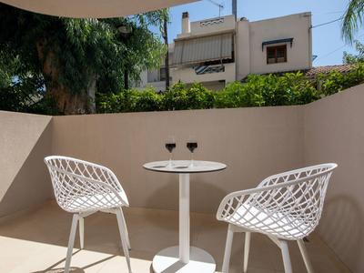 Two white chairs and a small table with two glasses on a sunny terrace next to a building facade.