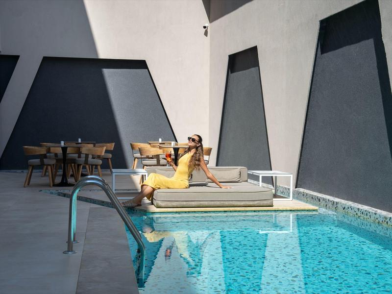 Woman relaxing on lounger beside clear blue pool with modern gray building in background.