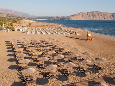 Sonnenschirme und Liegestühle in Reihen auf einem Sandstrand mit Blick auf das Meer und Berge im Hintergrund.