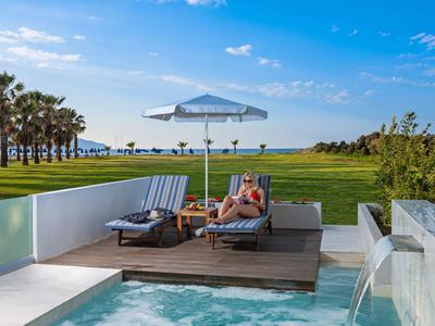 Luxuriöse Poolterrasse mit Liegestühlen und Sonnenschirm vor einem grünen Rasen und Blick aufs Meer.