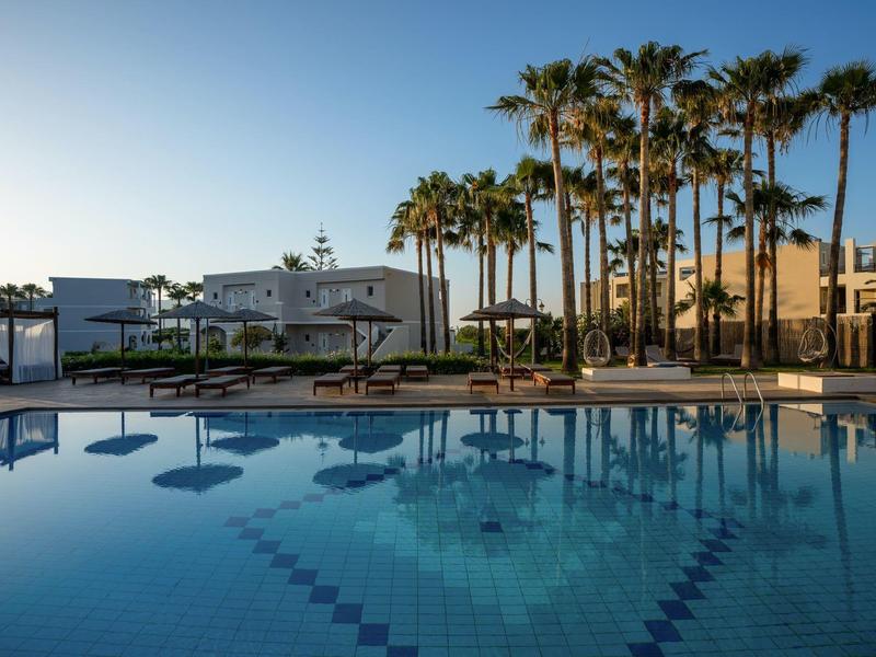 Hotelpool mit Palmen und Sonnenschirmen bei klarem Himmel und ruhiger Wasseroberfläche.